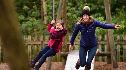 A girl in a pink jacket is being pushed on a zip slide by her mum. The adult is wearing a blue jacket and black bobble hat.